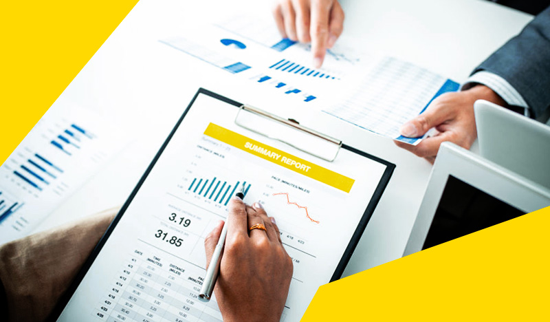 The image shows two people reviewing printed charts and graphs. One person is holding a clipboard with a summary report th...