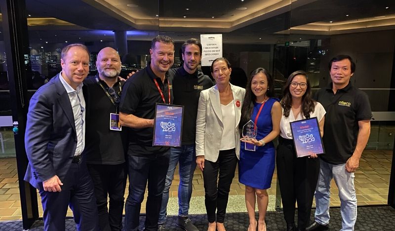 The image shows a group of eight people standing together indoors, possibly at an event or award ceremony. Three individua...