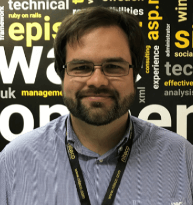 Man with glasses and beard wearing lanyard stands against a tech keyword wall
